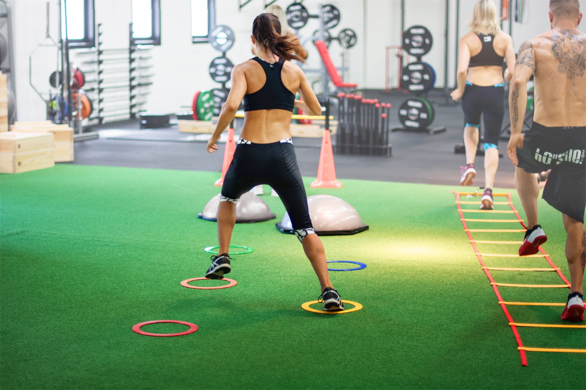 Athletes performing speed and agility footwork drills on a green commercial turf track with a speed ladder and agility training equipment.